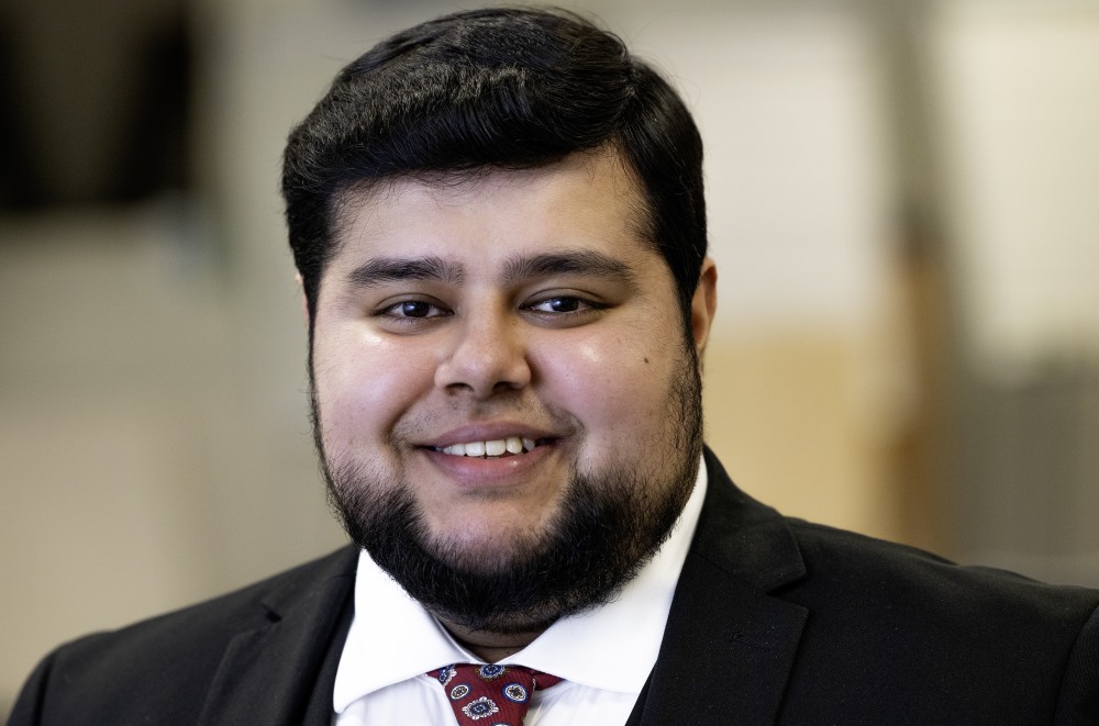 Photograph of Ibrahim, a smiling young man, wearing a black suit with a white shirt and red patterened tie.