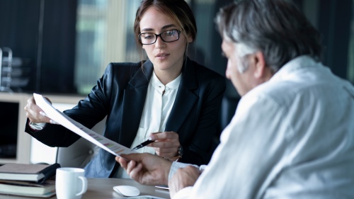 A woman dressed in business clothes uses a pen to point out something on a document to an older man sitting next to her