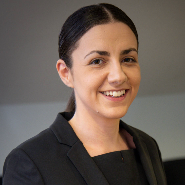 Carley is wearing a tailored dark suit, they have dark hair which is tied back.