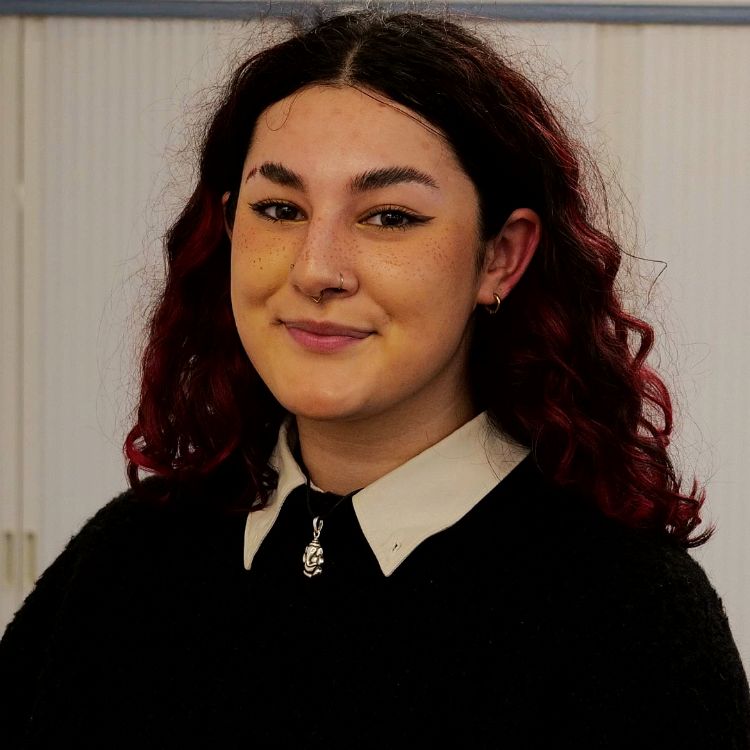Head and shoulder photo of Mia. Mia has curly shoulder length hair with redish/pink colours. Mia has nose piercings and wears a dark chunky jumper.
