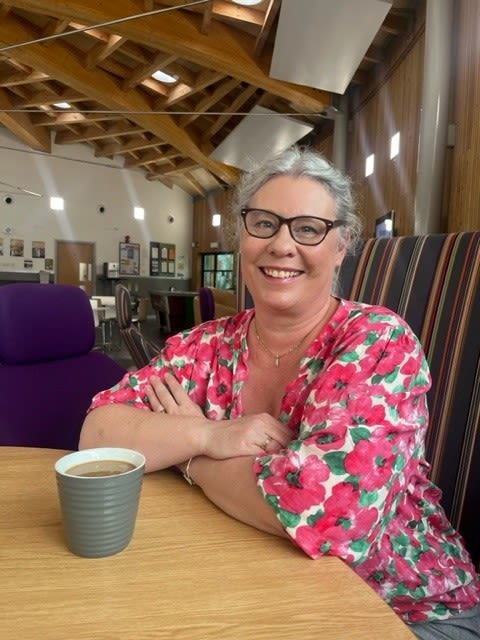 Photo of Helen, sat at a table with a mug of coffee, silver hair tied back with glasses and wearing a floral pink blouse.