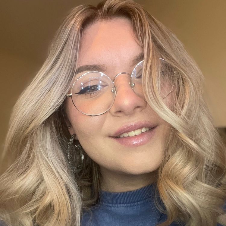 Head and shoulders photo of Charlotte. Charlotte has blonde mid length hair, wears round gold trimmed glasses and wears a blue jumper.