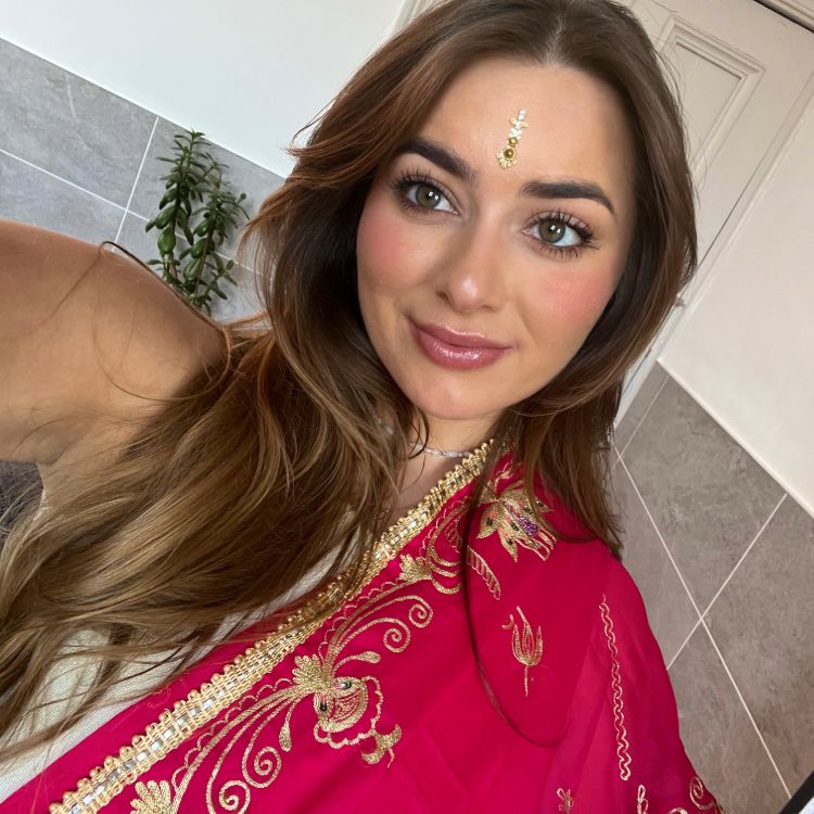 Head and shoulder photo of Priya. Priya has long mid brown highlighted hair and is wearing a red and gold embroidered sari.