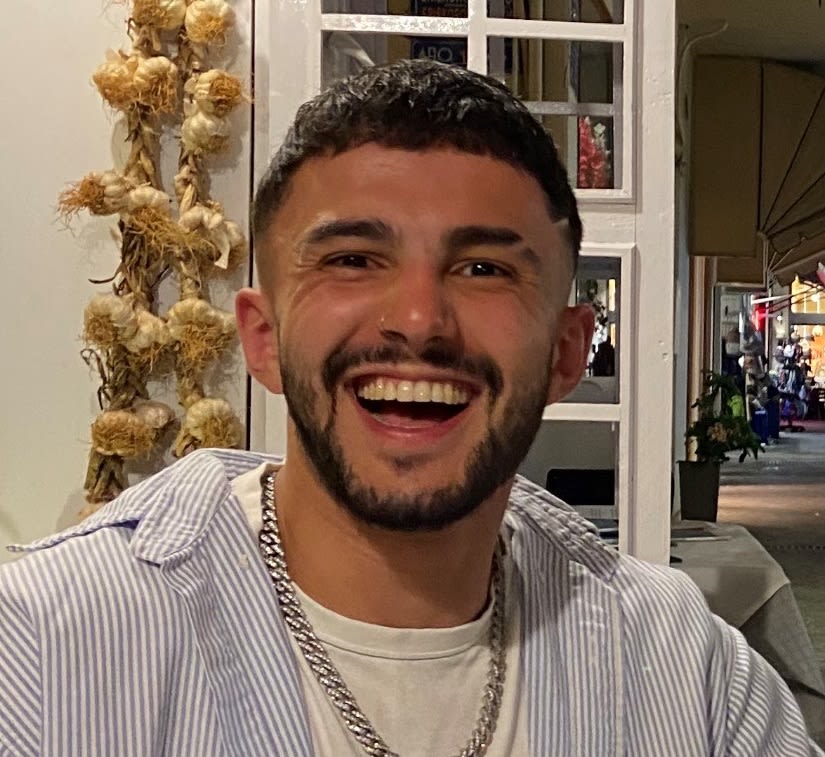 Photo of Samir, Adminstrative Officer. With short dark brown hair, a short beard and wearing a stripey blue shirt with white tshirt underneath.