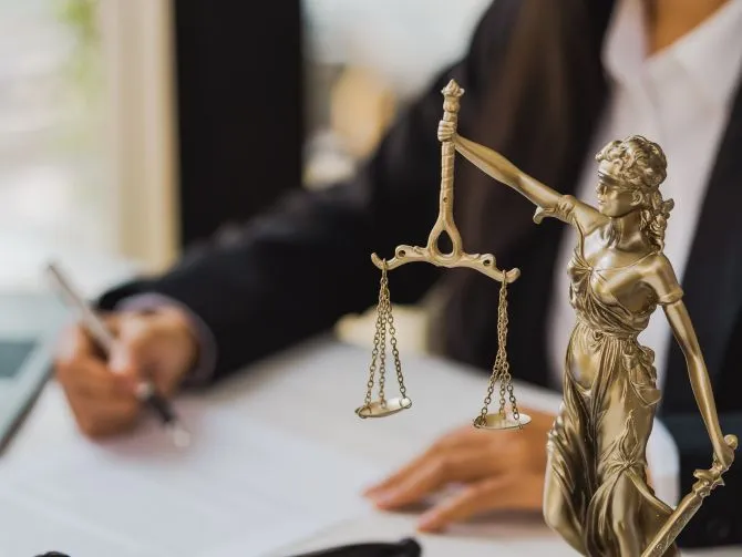 A person writing in a file in the background, with a small silver statue of Lady Justice in the foreground.