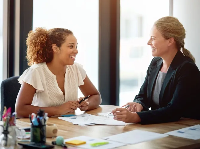 Two wwomen are shown at a meeting room table scattered with papers. They are smiling at each other, evidently in the middle of an animated discussion.