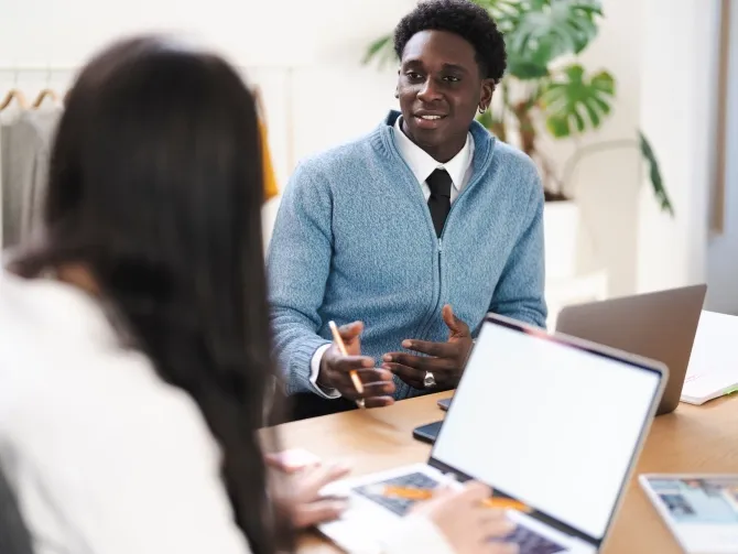 A young woman and young man are sitting at a desk, talking. The woman has a laptop open on the desk in front of her. the young man is smiling.