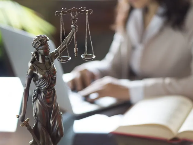 A view of a desktop with a statuette of Lady Justice standing on it. In the background, out of focus, a woman typing on a laptop can be seen.