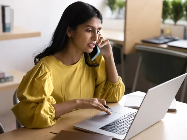 A young woman its at a desk, on her laptop while conducting a phone call on her mobile phone